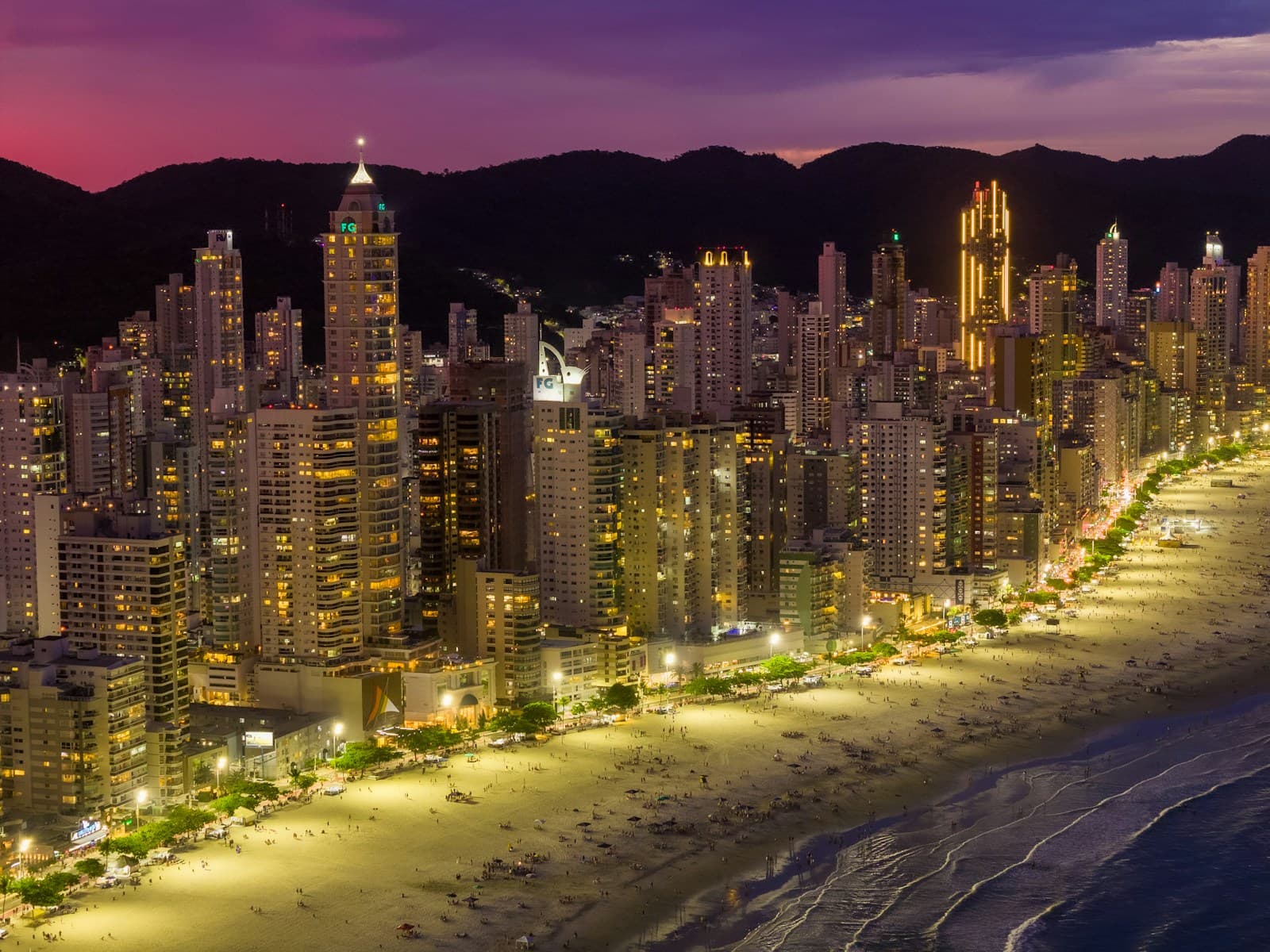 Skyline de Balneário Camboriú visto da Barra Sul, com as torres da praia central iluminadas ao entardecer e os morros ao fundo